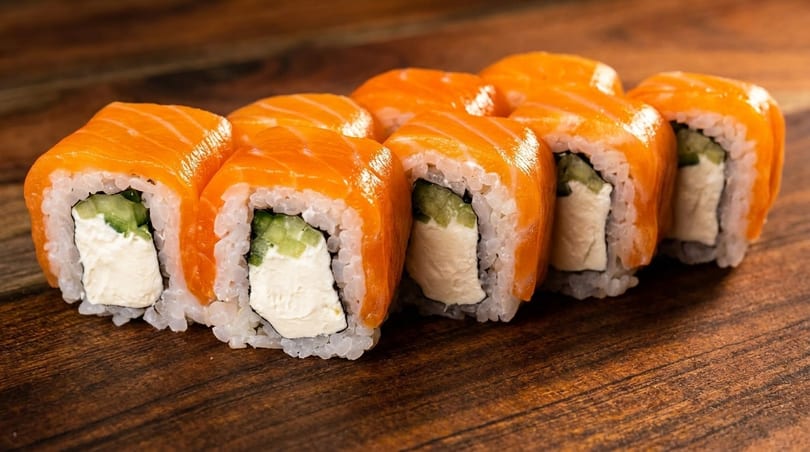Six salmon sushi rolls with cream cheese and cucumber on a wooden surface.