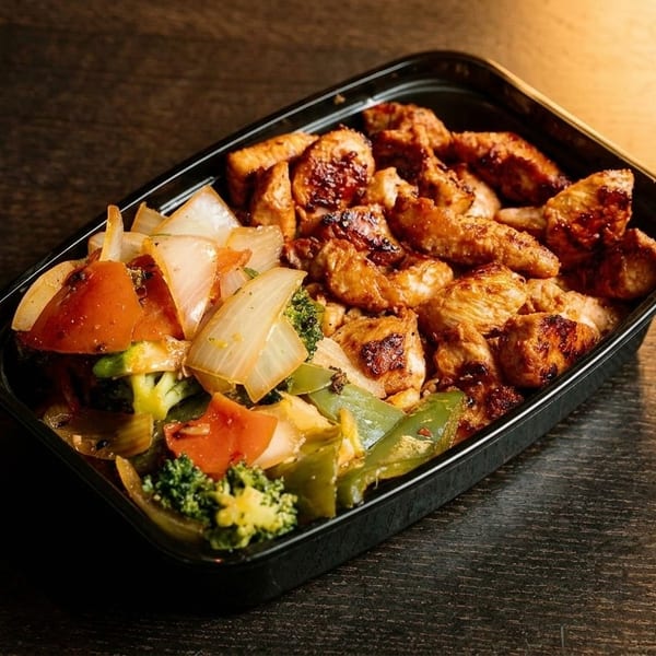 Grilled chicken and stir-fried vegetables in a black takeout container on a dark wooden table.