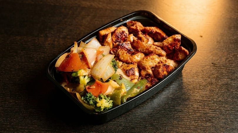 Grilled chicken pieces and mixed vegetables, including broccoli, onions, and bell peppers, in a black container.