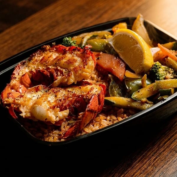 Grilled lobster tails with rice, mixed vegetables, and a lemon wedge in a black container.