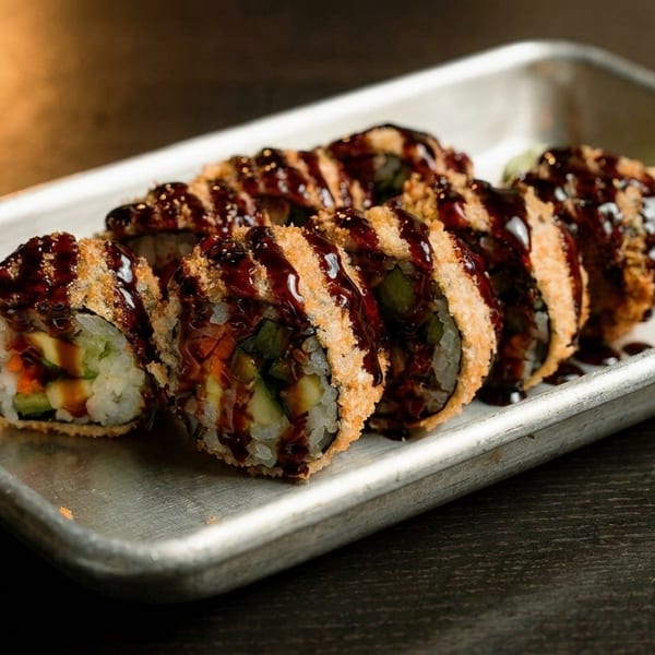 Crispy fried sushi rolls drizzled with dark sauce, served on a metal tray with pickled ginger.